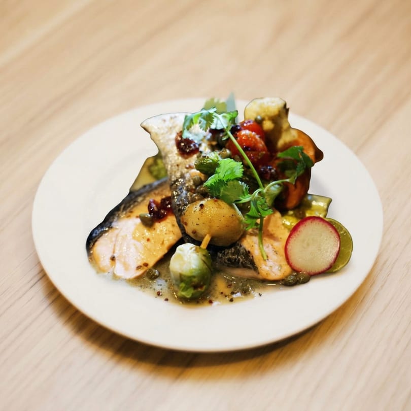 Grilled salmon with roasted vegetables, capers, and fresh herbs on a white plate.