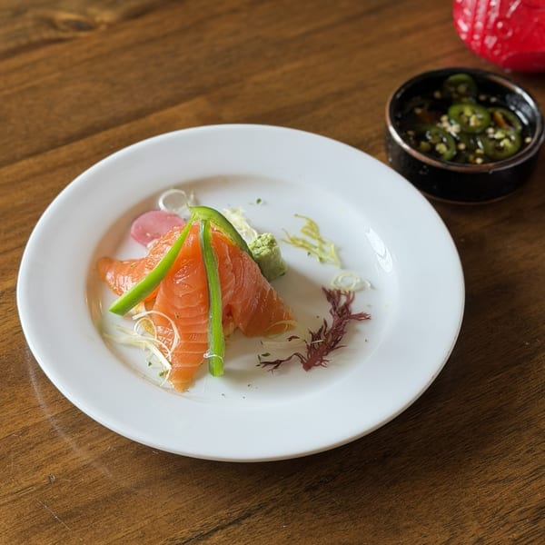 Salmon sashimi with green peppers, wasabi, and seaweed on a white plate with a side of jalapeño sauce.